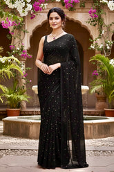 Woman in a black saree with sequins standing in an outdoor setting with plants and flowers.
