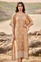 Woman wearing a beige kaftan with white patterns standing in a natural setting. Product by Umaid Couture.