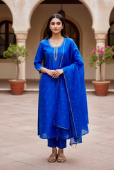 Woman in a blue traditional outfit standing in an outdoor setting with arches and plants.