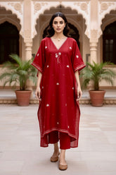 Woman wearing a red Kaftan with white patterns in an outdoor setting with architectural elements and plants.