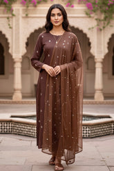 Woman in a brown traditional kurta standing in front of an architectural background with arches and plants. Product by Umaid Couture