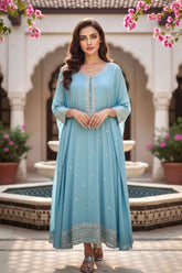 Woman in a light blue Kaftan standing in front of a decorative fountain with pink flowers. Product by Umaid Couture