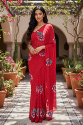 An Indian woman standing in a garden courtyard wearing a red chiffon saree embellished with stylised peacock motifs made with resham and silver, golden sequins by Umaid Couture.