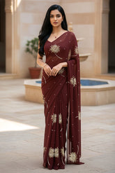 An Indian woman standing in a hotel courtyard wearing a coco brown chiffon saree embellished with shevanti flower motifs made with shimmering Zari, Sequins, and Aari-Zardosi craft by Umaid Couture.