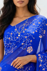 An Indian woman standing in a minimal white courtyard wearing a royal blue chiffon saree embellished with vgolden sliver and pink sequin vines of Stylized Jasmine by Umaid Couture.