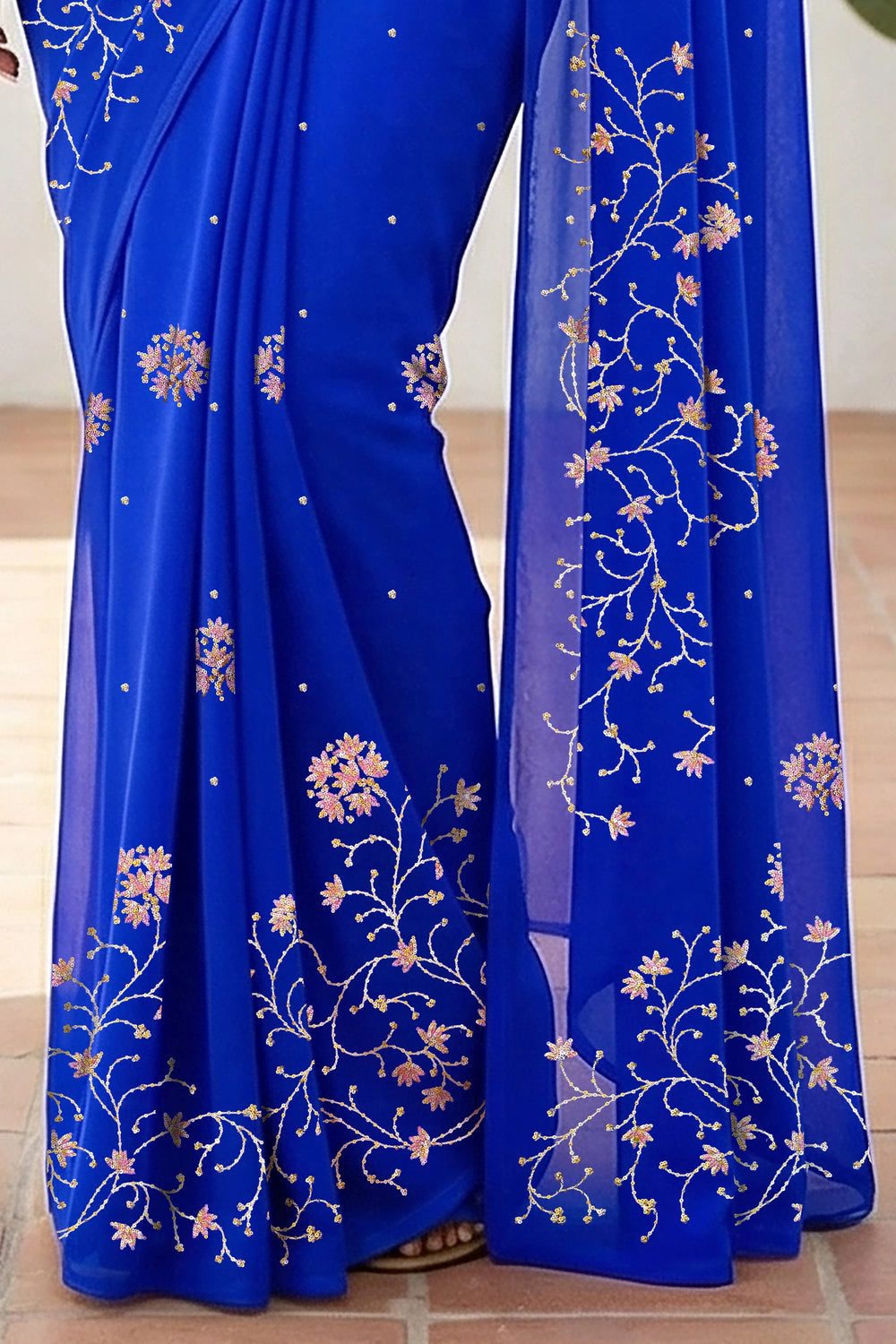 An Indian woman standing in a minimal white courtyard wearing a royal blue chiffon saree embellished with vgolden sliver and pink sequin vines of Stylized Jasmine by Umaid Couture.