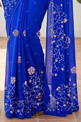 An Indian woman standing in a minimal white courtyard wearing a royal blue chiffon saree embellished with vgolden sliver and pink sequin vines of Stylized Jasmine by Umaid Couture.