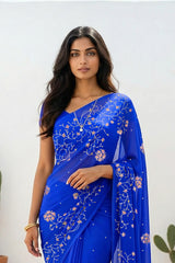 An Indian woman standing in a minimal white courtyard wearing a royal blue chiffon saree embellished with vgolden sliver and pink sequin vines of Stylized Jasmine by Umaid Couture.
