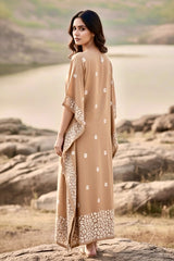 Woman in a beige kaftan with white floral patterns standing on rocky terrain near a body of water. Product by Umaid Couture.