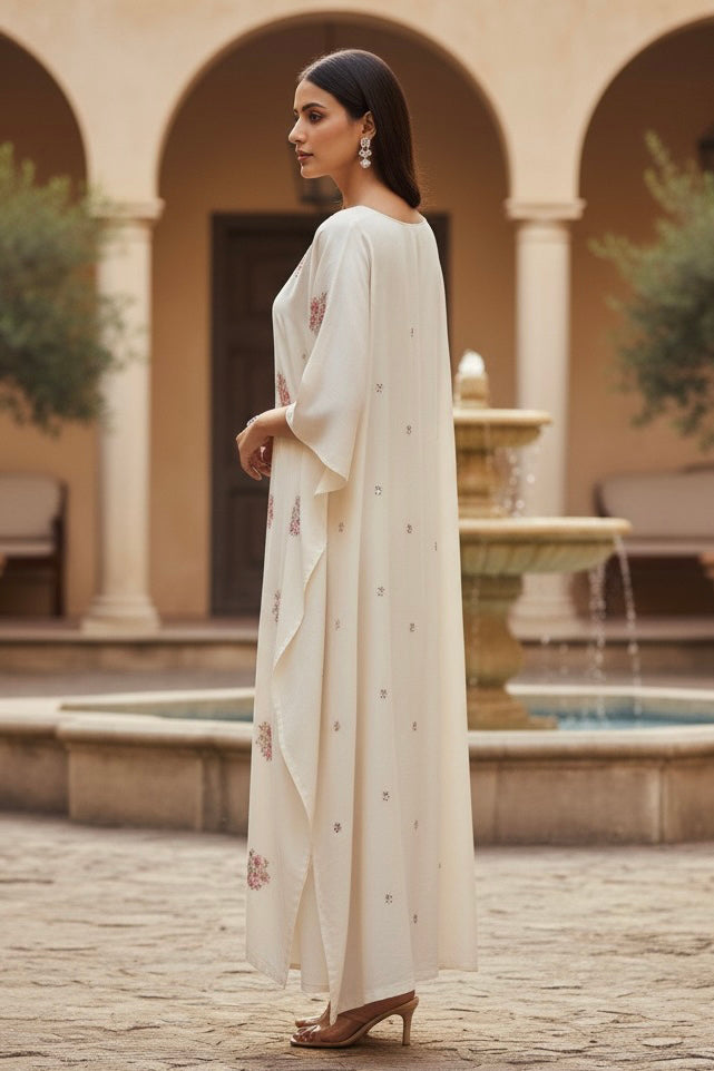 Woman in a long, light-colored kaftan standing in an outdoor setting with arches and a fountain. Product by Umaid Couture
