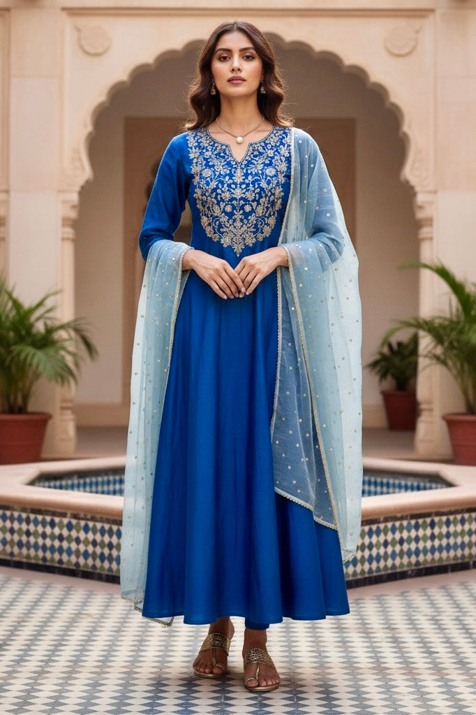 Woman in a blue traditional outfit with intricate patterns standing in a decorative outdoor setting. Product by Umaid Couture