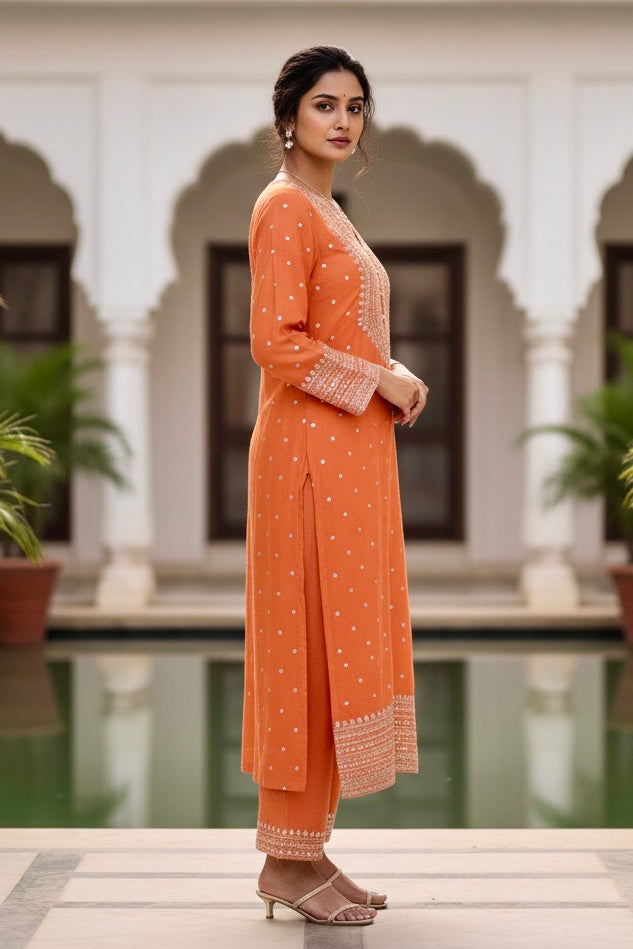 Woman in an orange traditional kurta standing in front of a pool with architectural elements in the background. Product by Umaid Couture