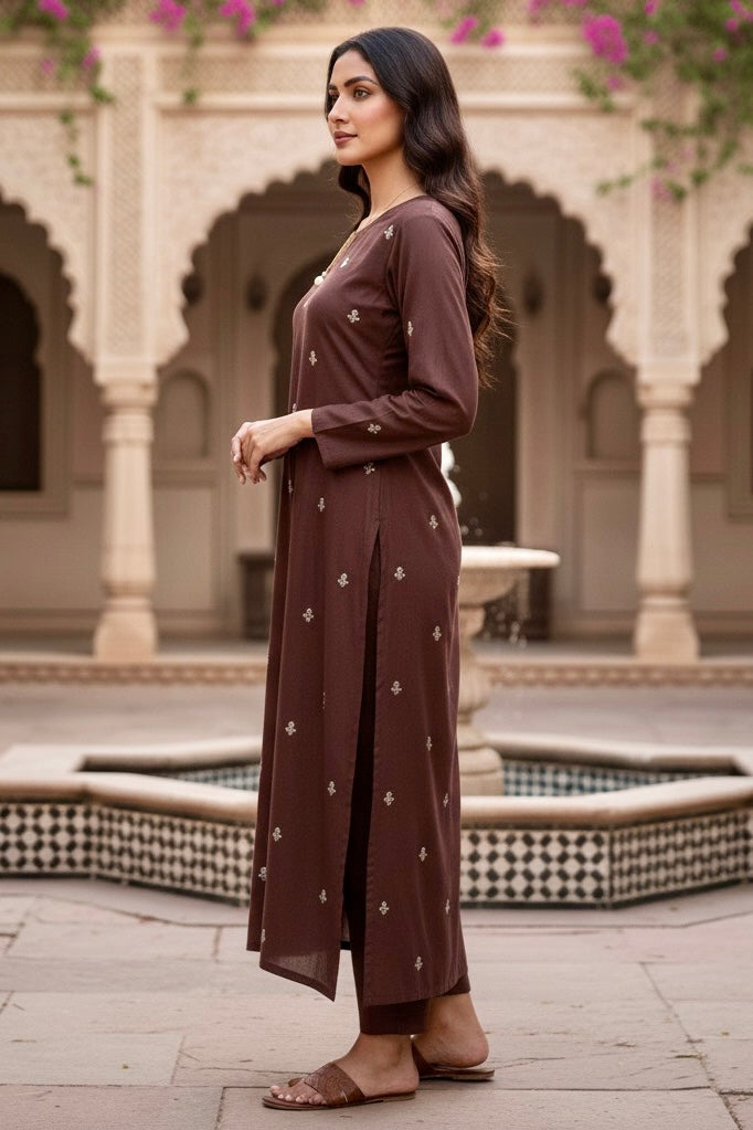 Woman in a brown kurta standing in an outdoor setting with architectural elements. Product by Umaid Couture