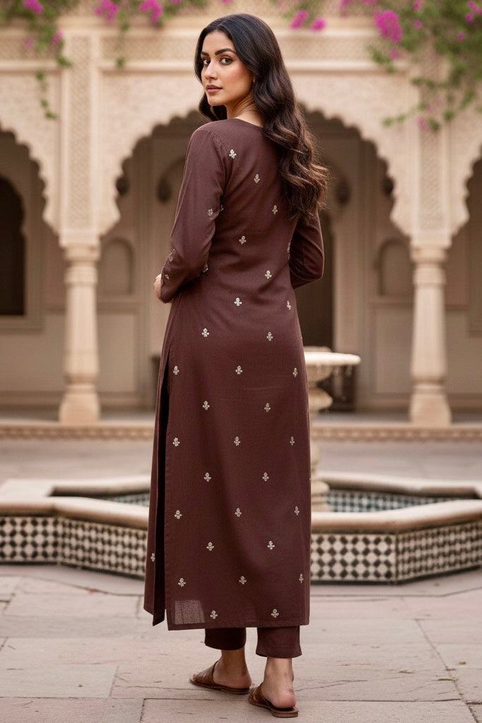 Woman in a brown traditional outfit standing in a courtyard with arches and floral decorations. Product by Umaid Couture