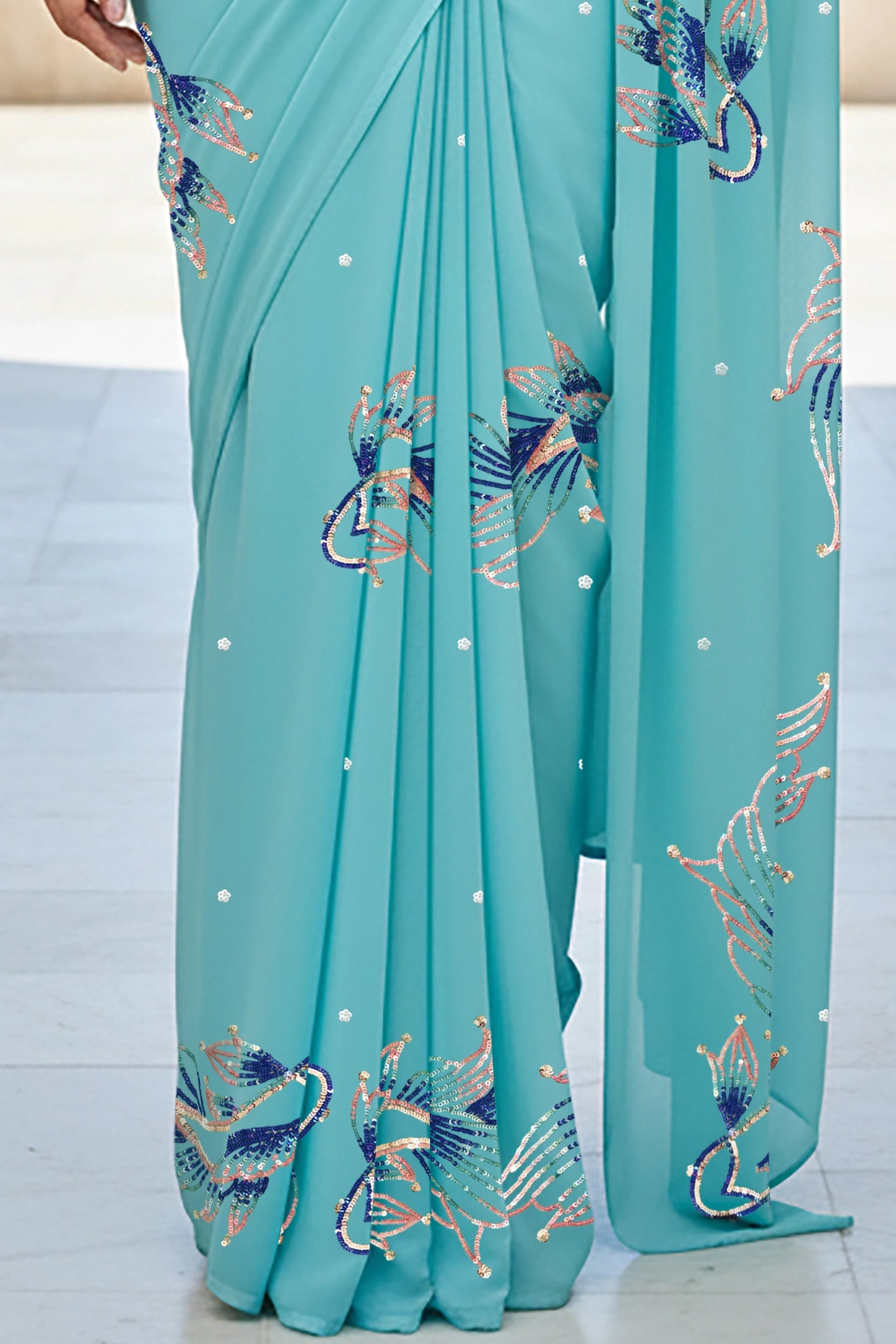 An Indian woman standing in a seaside hotel courtyard wearing sea green color chiffon saree embellished with blue, pink, cyan and sliver sequin of fish motifs by Umaid Couture.  
