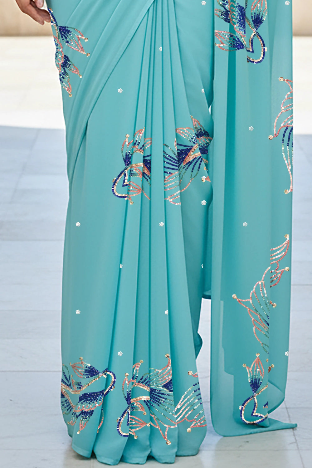 An Indian woman standing in a seaside hotel courtyard wearing sea green color chiffon saree embellished with blue, pink, cyan and sliver sequin of fish motifs by Umaid Couture.  

