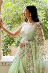 Woman in a light green saree with floral patterns standing in front of a window with greenery outside.