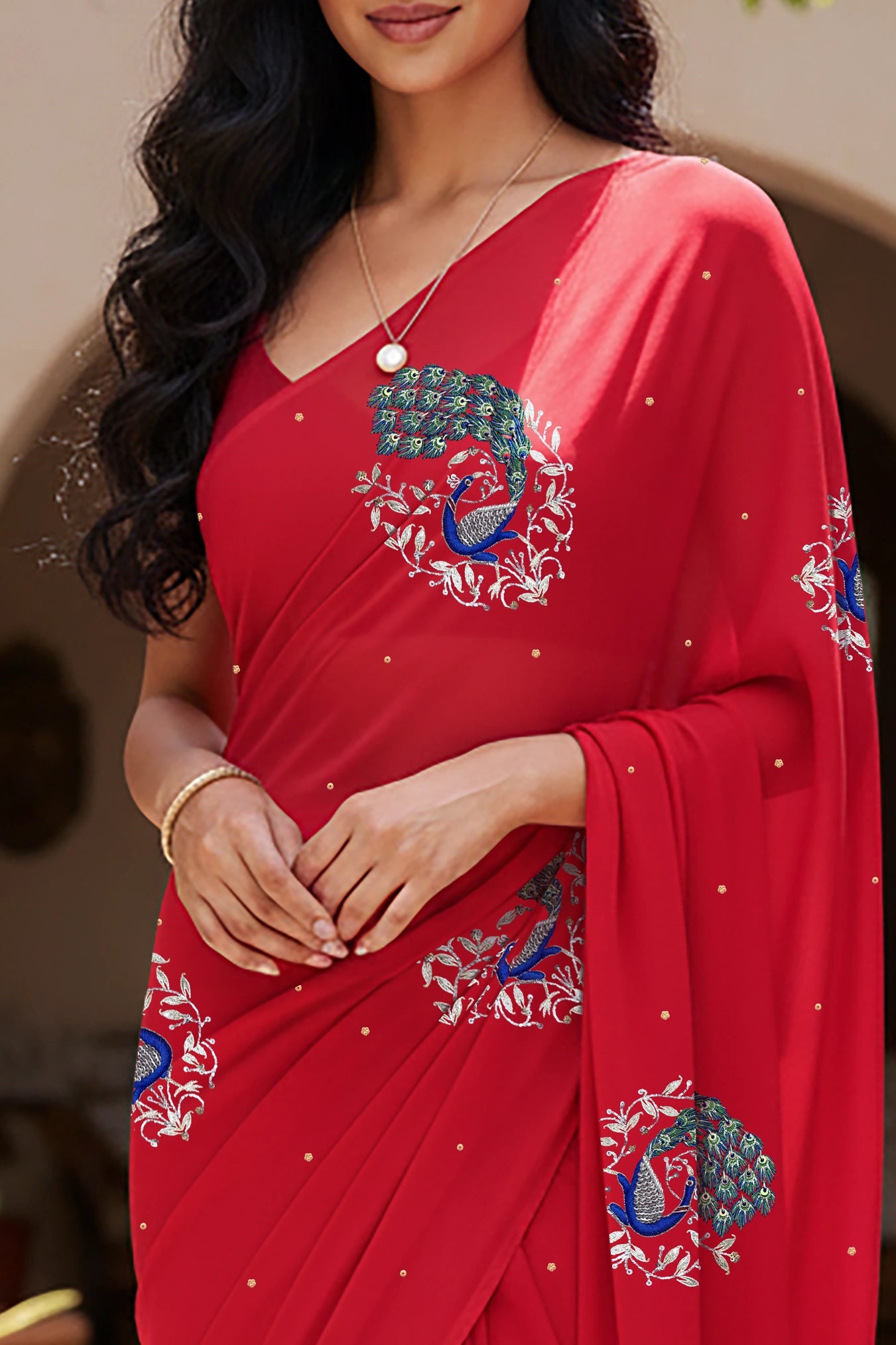 An Indian woman standing in a garden courtyard wearing a tomato red chiffon saree embellished with blue, sliver and green sequin of peacock motifs by Umaid Couture.  
