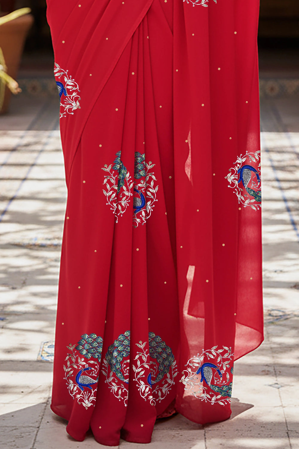 An Indian woman standing in a garden courtyard wearing a tomato red chiffon saree embellished with blue, sliver and green sequin of peacock motifs by Umaid Couture.  
