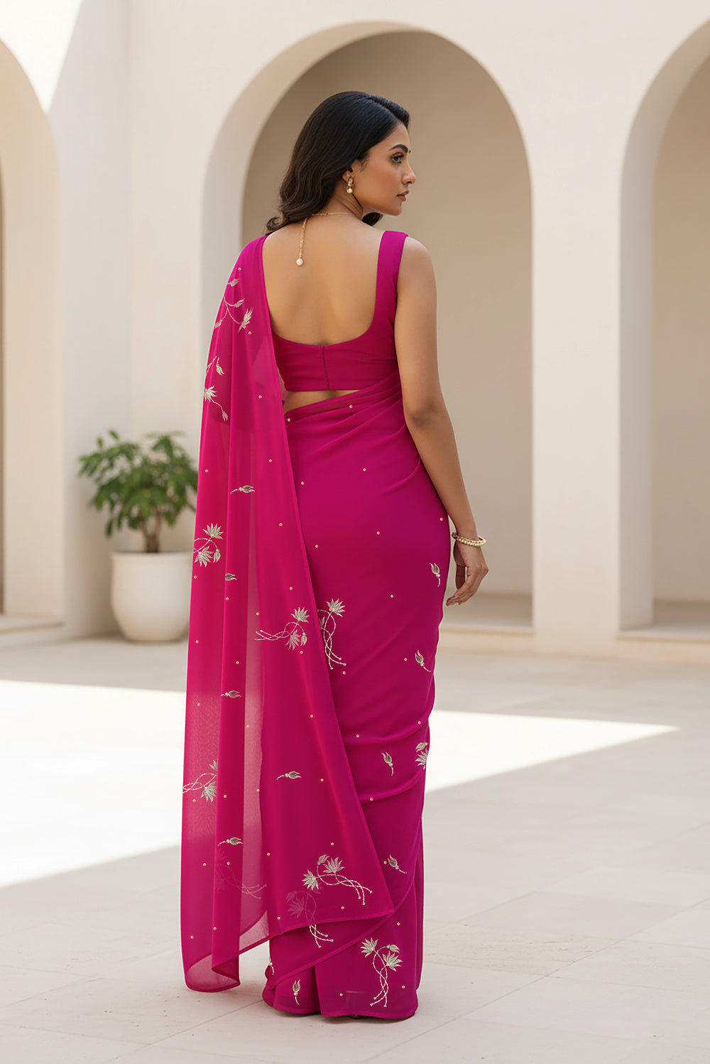 Woman in a pink saree with a white floral pattern standing in a sunlit room with arches. Product By Umaid Couture