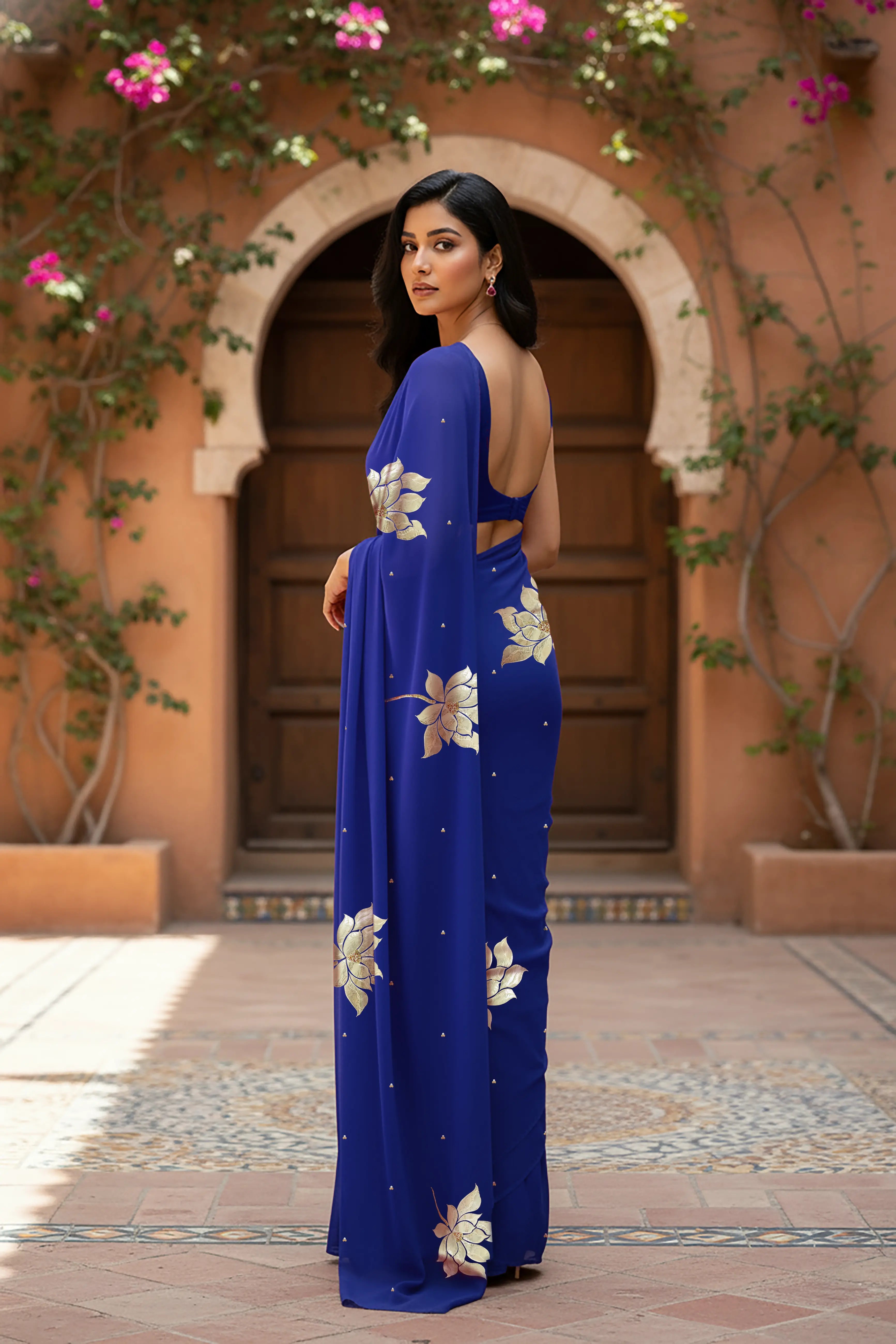 An Indian woman standing in a garden courtyard wearing blue chiffon saree embellished with large gota lotus flower motifs by Umaid Couture.  
