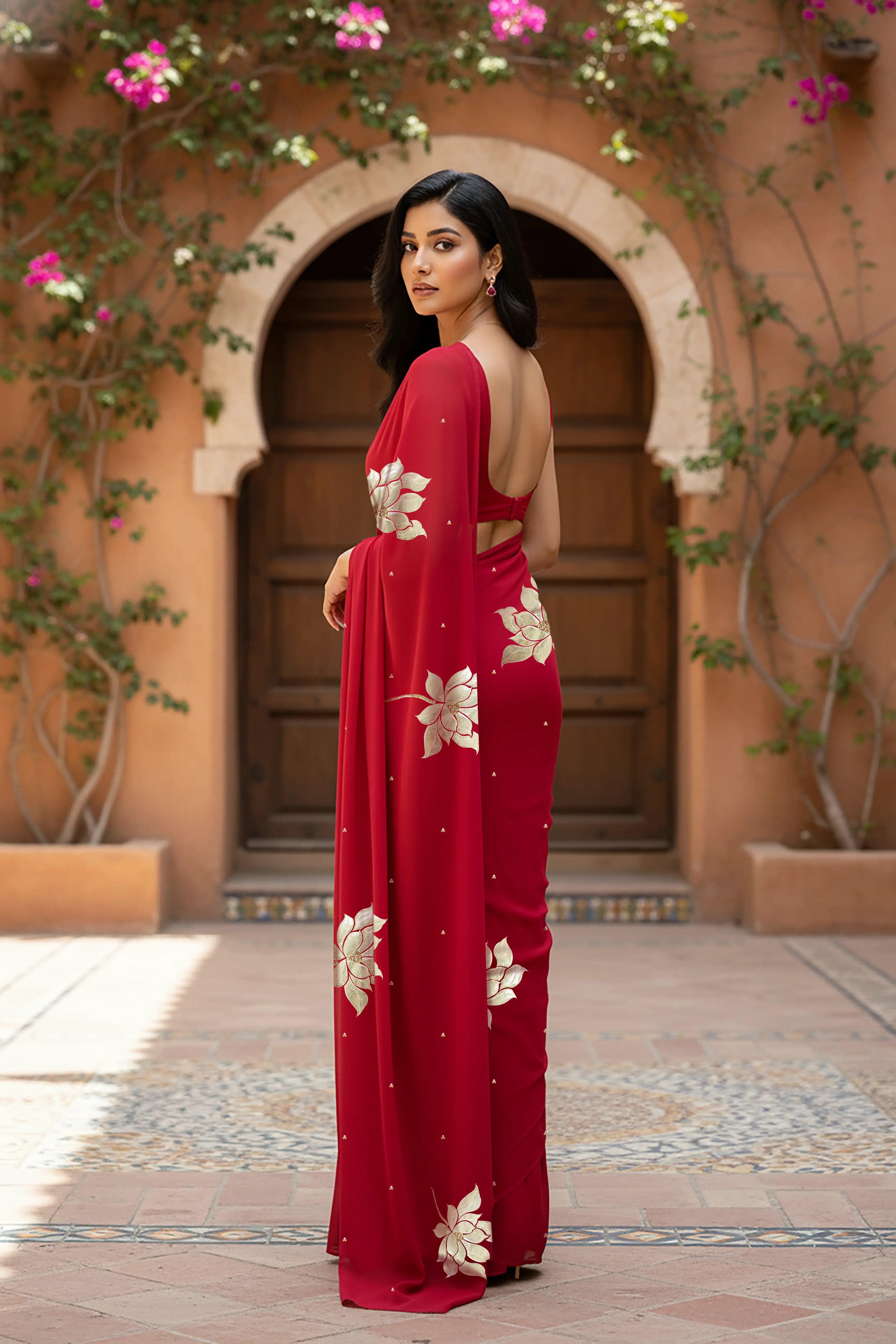 An Indian woman standing in a garden courtyard wearing red chiffon saree embellished with large gota lotus flower motifs by Umaid Couture.  
