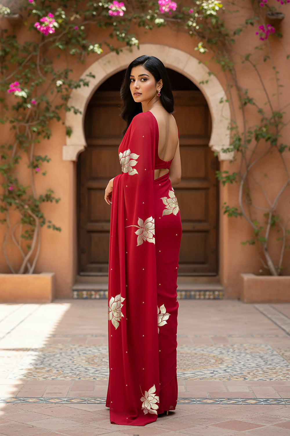 An Indian woman standing in a garden courtyard wearing red chiffon saree embellished with large gota lotus flower motifs by Umaid Couture.  
