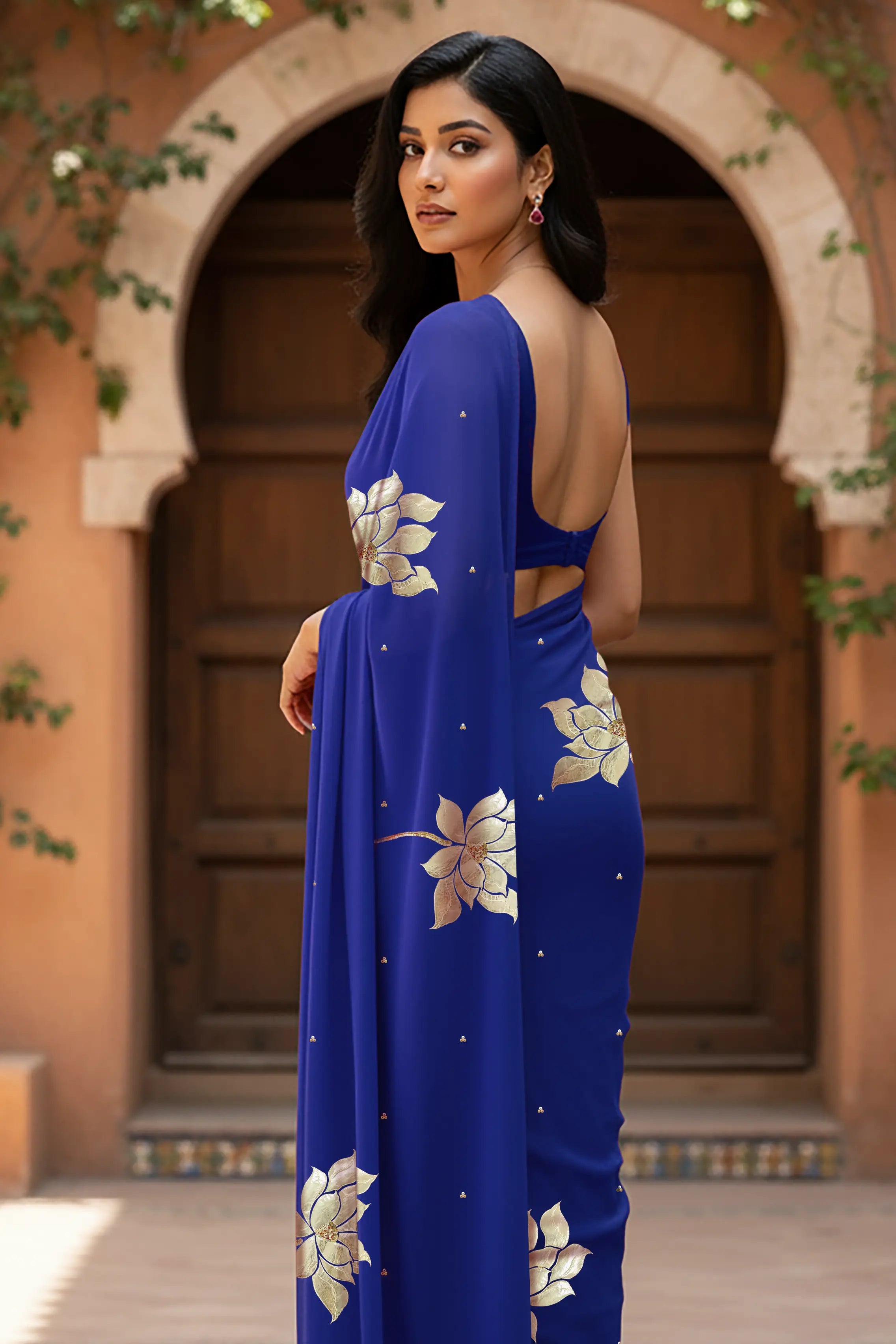 An Indian woman standing in a garden courtyard wearing blue chiffon saree embellished with large gota lotus flower motifs by Umaid Couture.  
