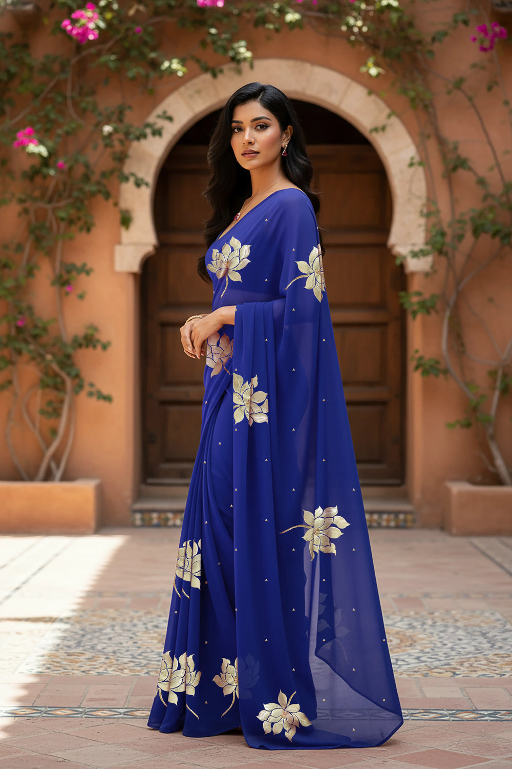 An Indian woman standing in a garden courtyard wearing blue chiffon saree embellished with large gota lotus flower motifs by Umaid Couture.  
