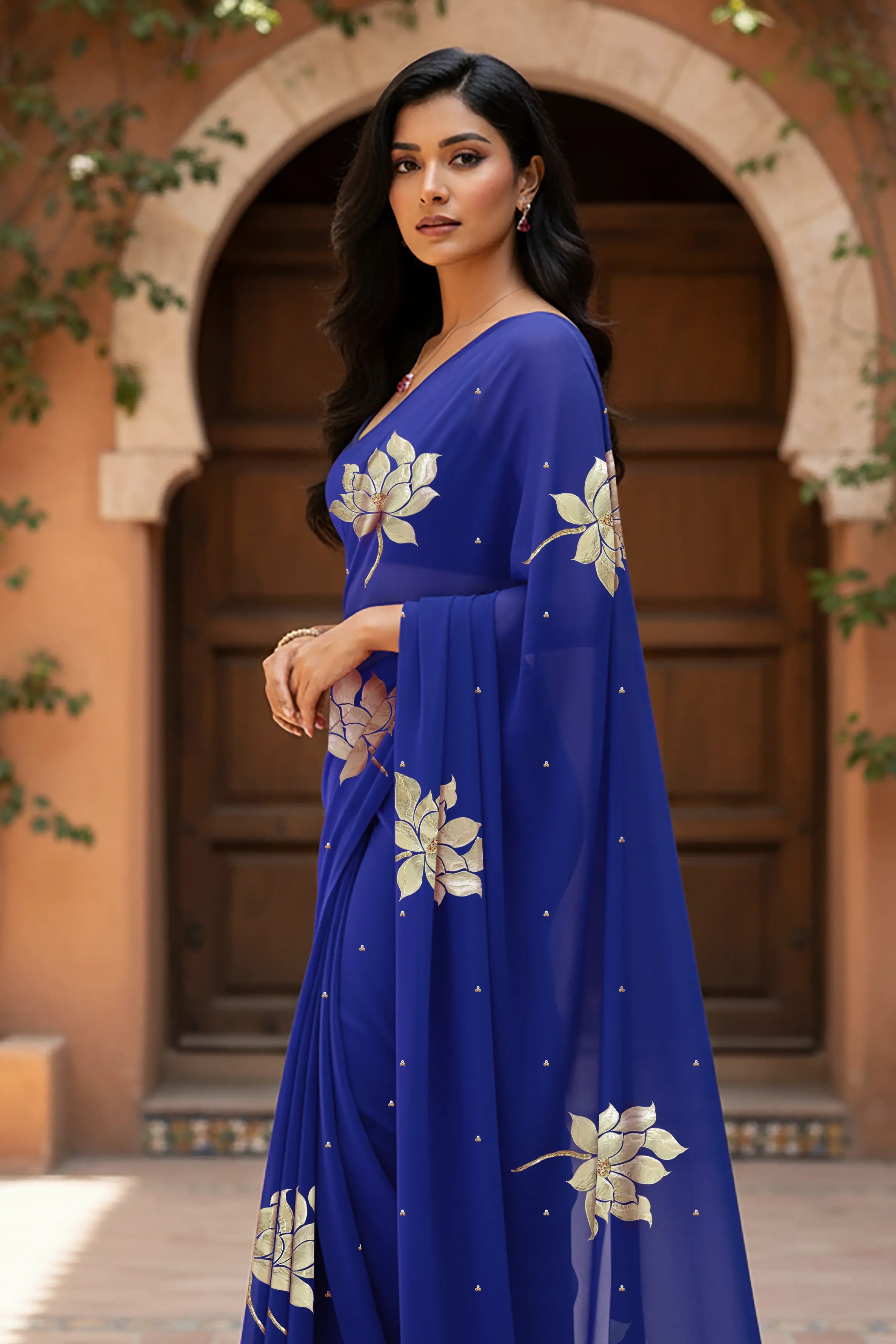 An Indian woman standing in a garden courtyard wearing blue chiffon saree embellished with large gota lotus flower motifs by Umaid Couture.  
