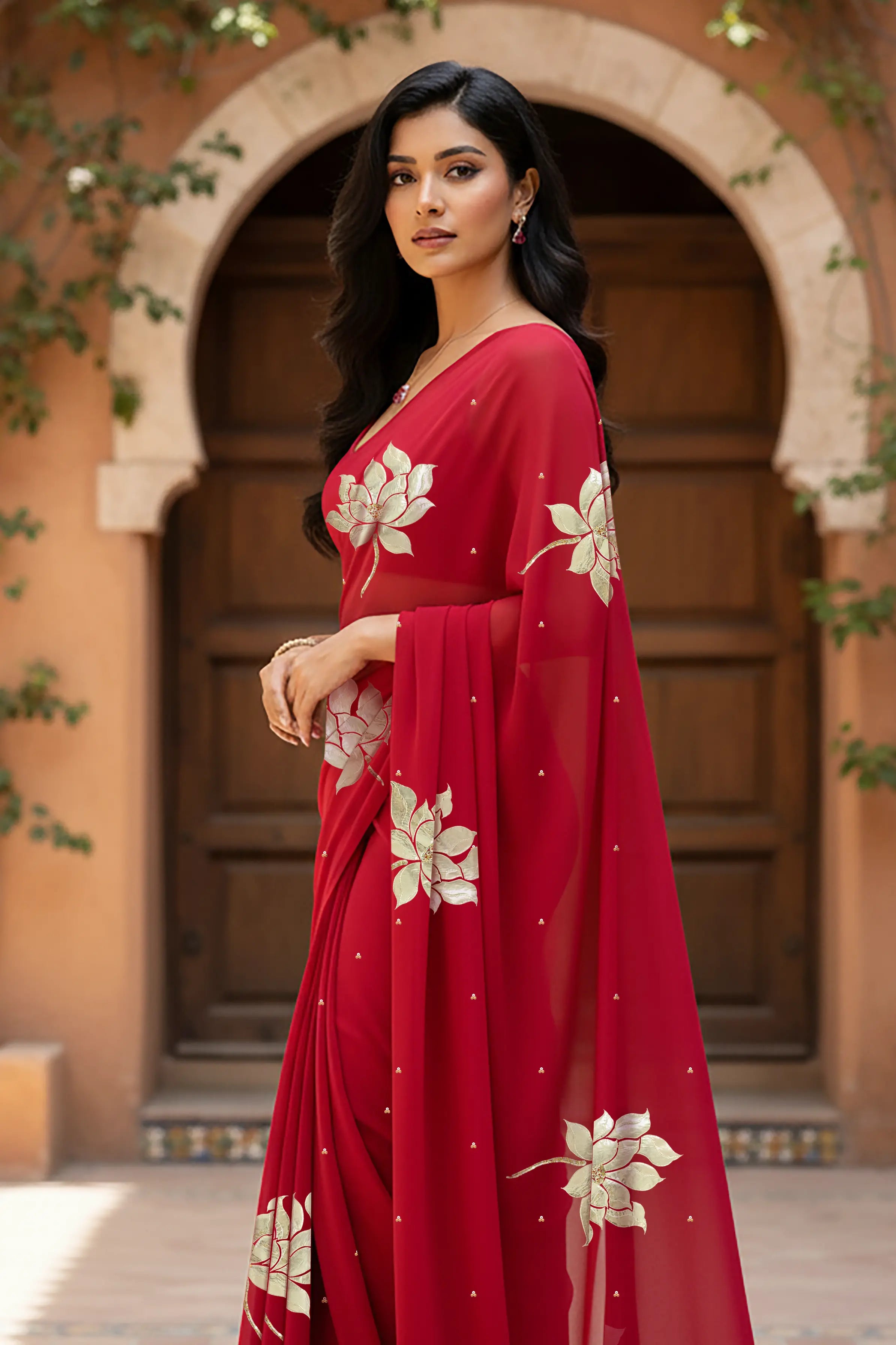 An Indian woman standing in a garden courtyard wearing red chiffon saree embellished with large gota lotus flower motifs by Umaid Couture.  
