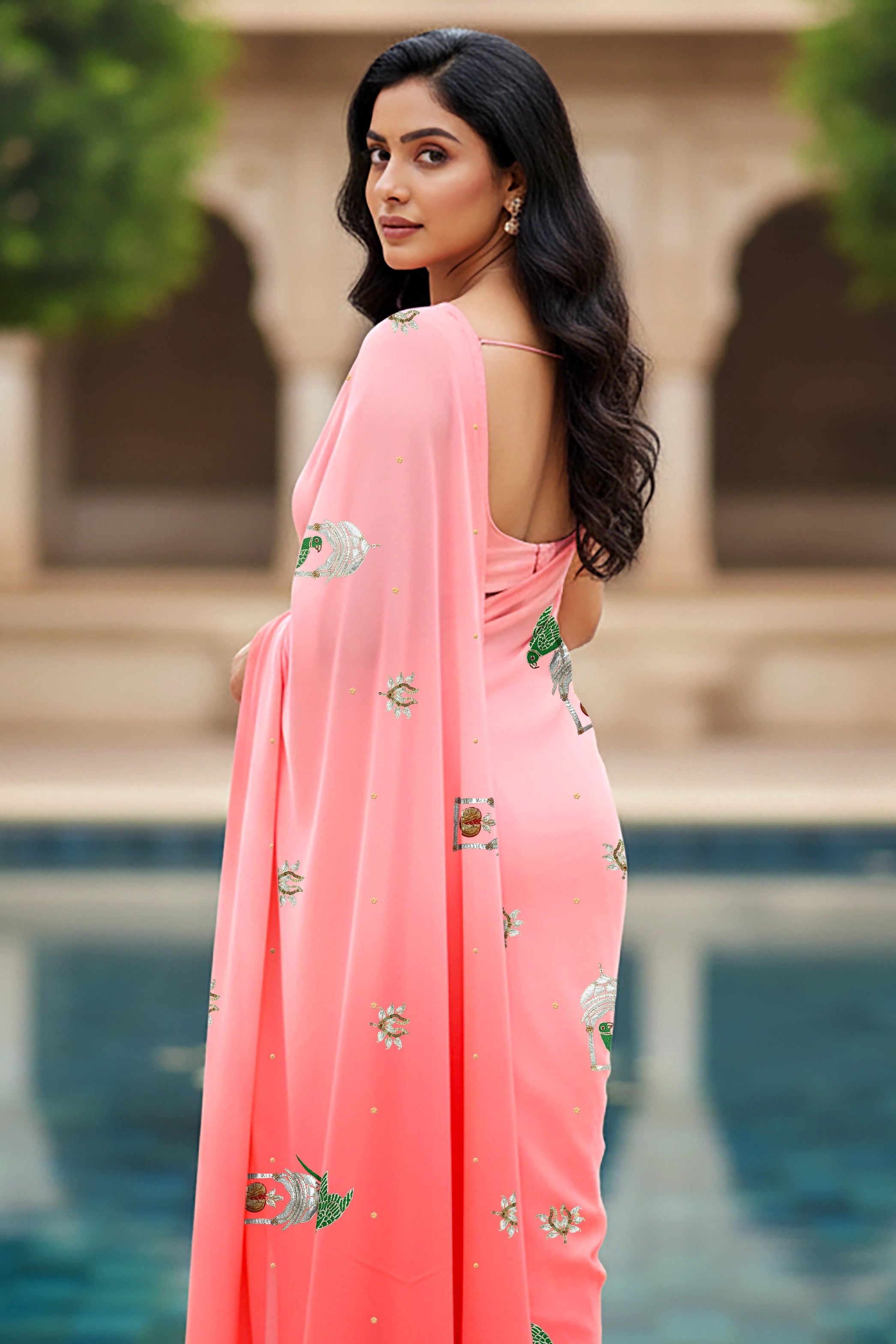 An Indian woman standing in a garden courtyard wearing a gradient of pink chiffon saree embellished with green and sliver sequin of l parrot siting in heritage style window motifs by Umaid Couture.  
