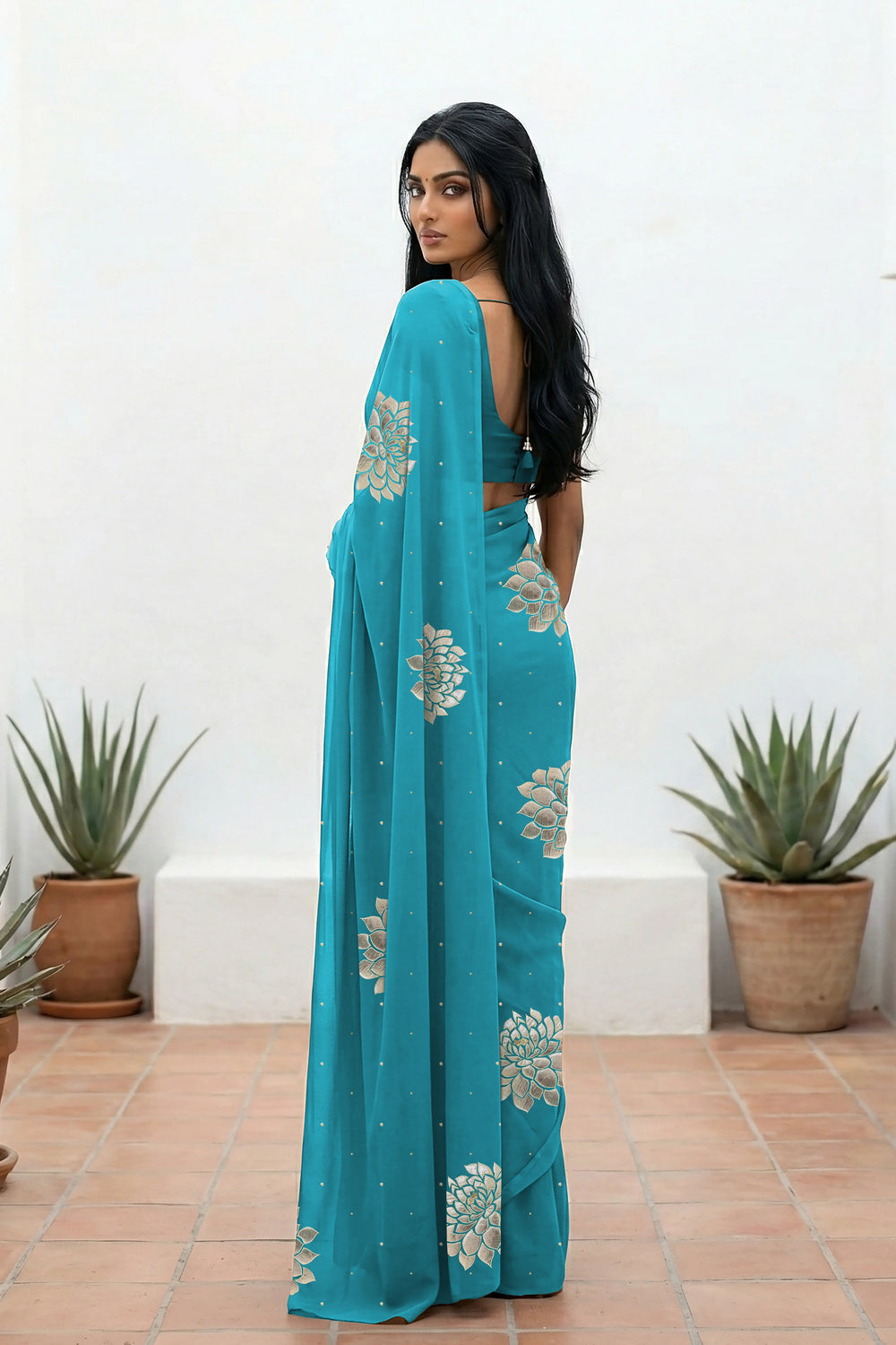 An Indian woman standing in a minimal white courtyard wearing a middle blue chiffon saree embellished with golden and sliver sequin of Dahlia flower motifs by Umaid Couture.  
