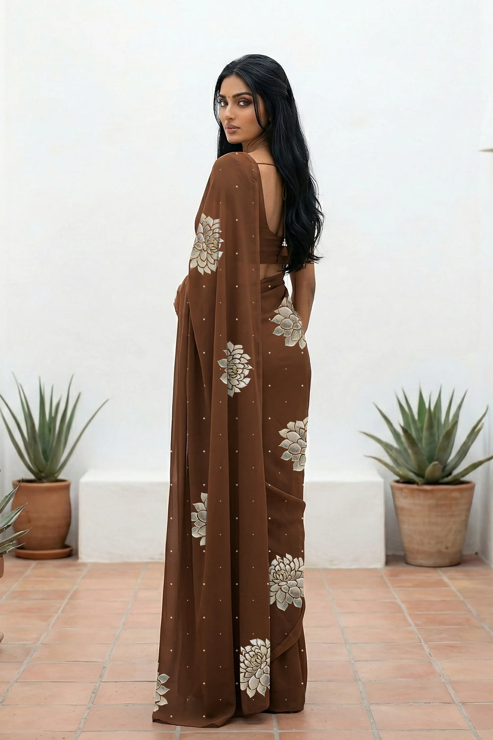 An Indian woman standing in a minimal white courtyard wearing a chocolate brown chiffon saree embellished with golden and sliver sequin of Dahlia flower motifs by Umaid Couture.  
