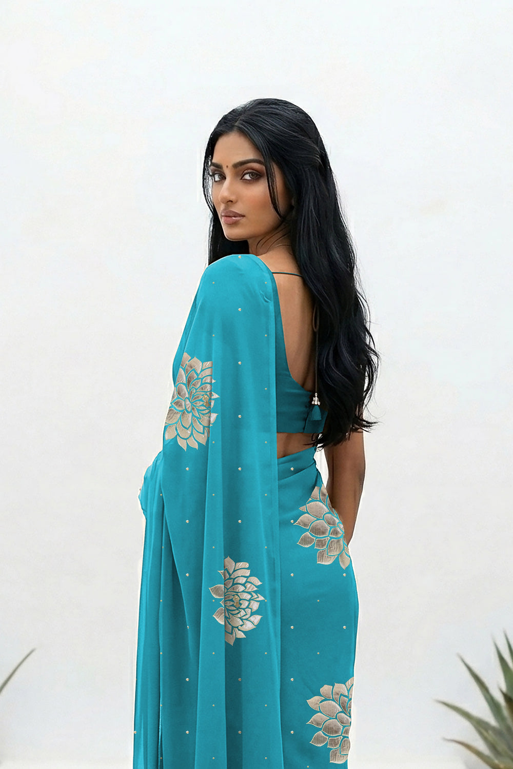 An Indian woman standing in a minimal white courtyard wearing a middle blue chiffon saree embellished with golden and sliver sequin of Dahlia flower motifs by Umaid Couture.  
