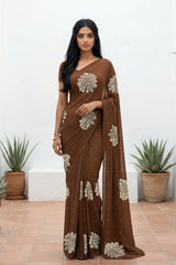 An Indian woman standing in a minimal white courtyard wearing a chocolate brown chiffon saree embellished with golden and sliver sequin of Dahlia flower motifs by Umaid Couture.  
