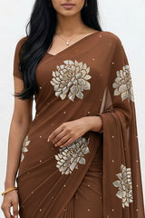 An Indian woman standing in a minimal white courtyard wearing a chocolate brown chiffon saree embellished with golden and sliver sequin of Dahlia flower motifs by Umaid Couture.  
