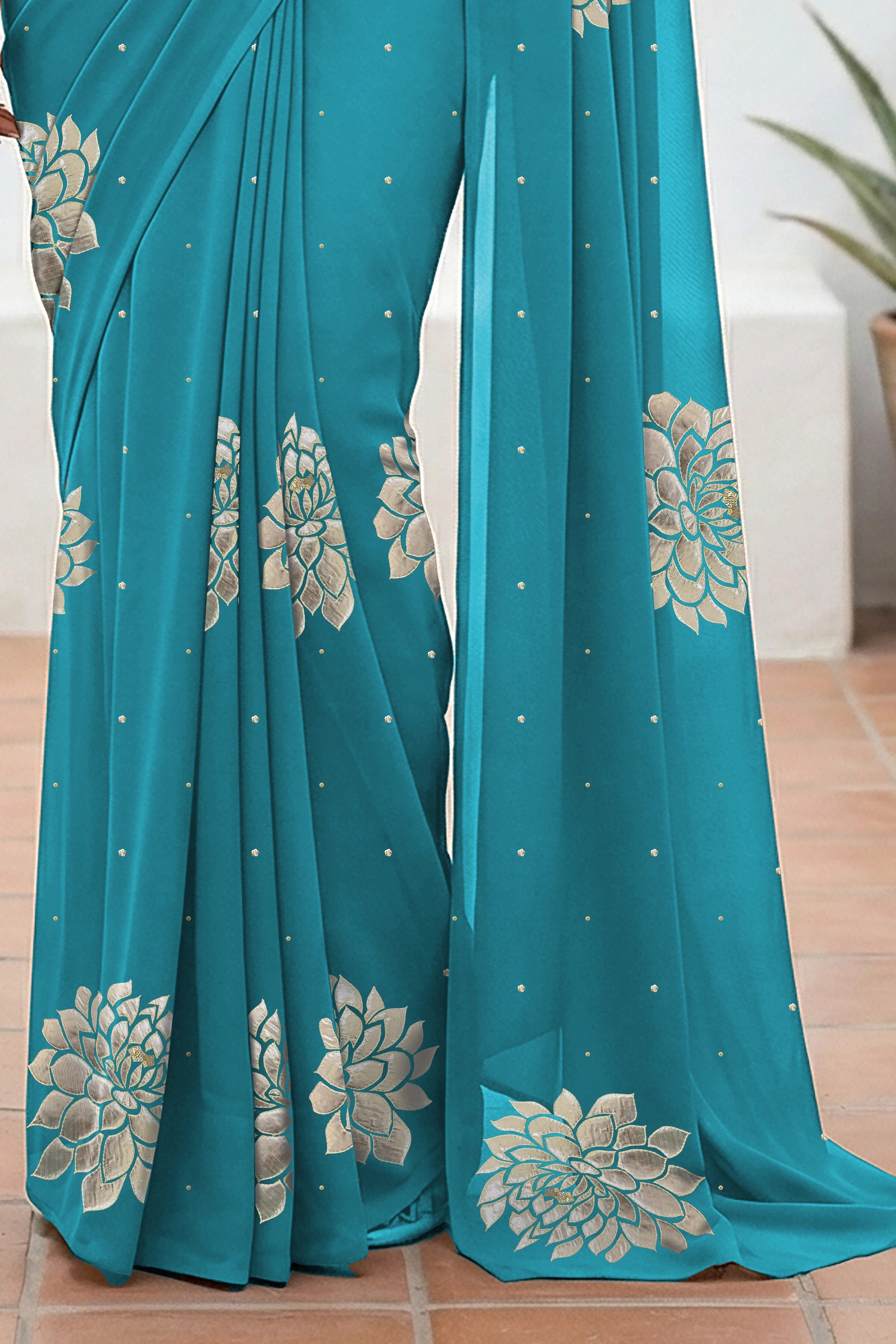 An Indian woman standing in a minimal white courtyard wearing a middle blue chiffon saree embellished with golden and sliver sequin of Dahlia flower motifs by Umaid Couture.  
