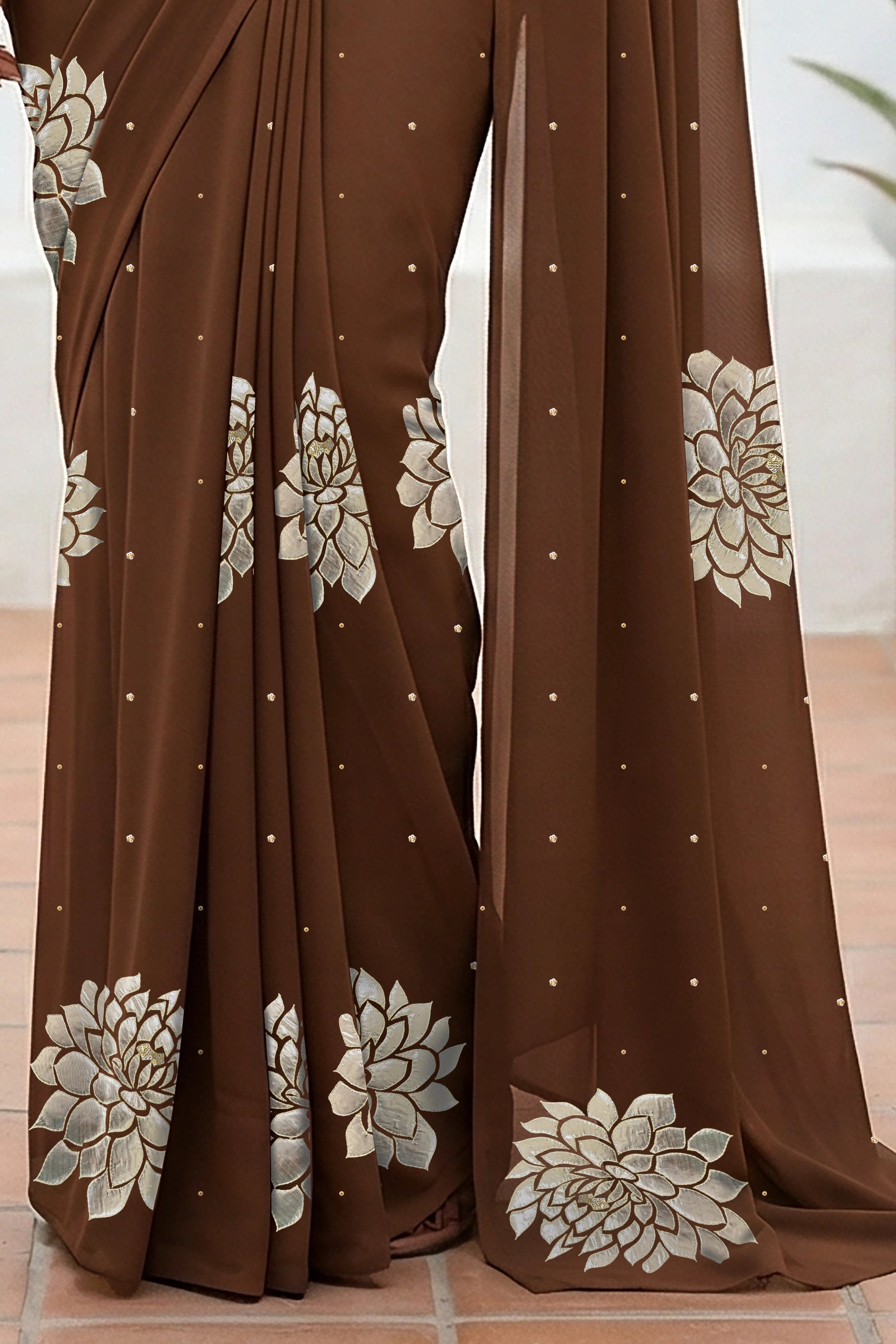 An Indian woman standing in a minimal white courtyard wearing a chocolate brown chiffon saree embellished with golden and sliver sequin of Dahlia flower motifs by Umaid Couture.  
