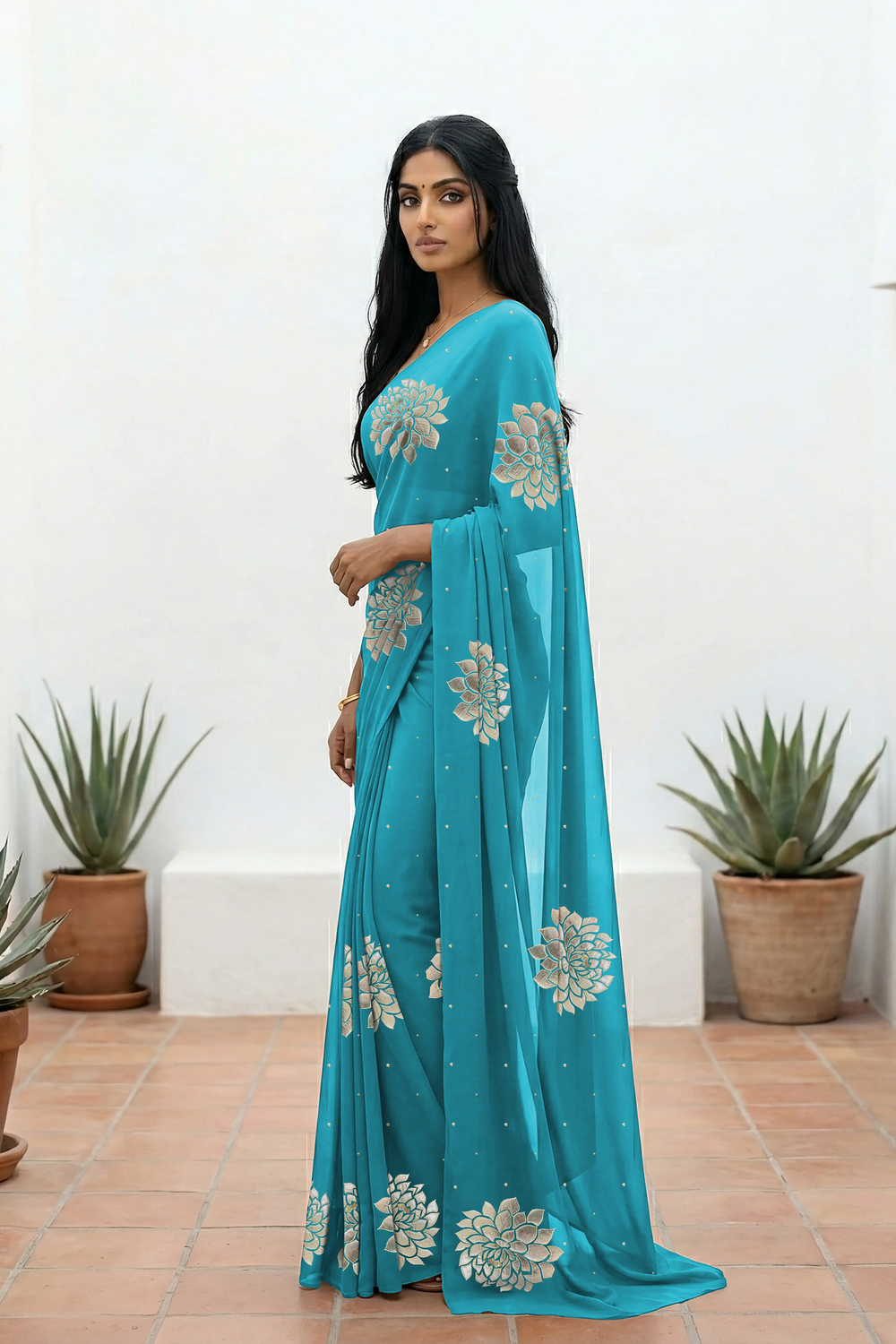 An Indian woman standing in a minimal white courtyard wearing a middle blue chiffon saree embellished with golden and sliver sequin of Dahlia flower motifs by Umaid Couture.  
