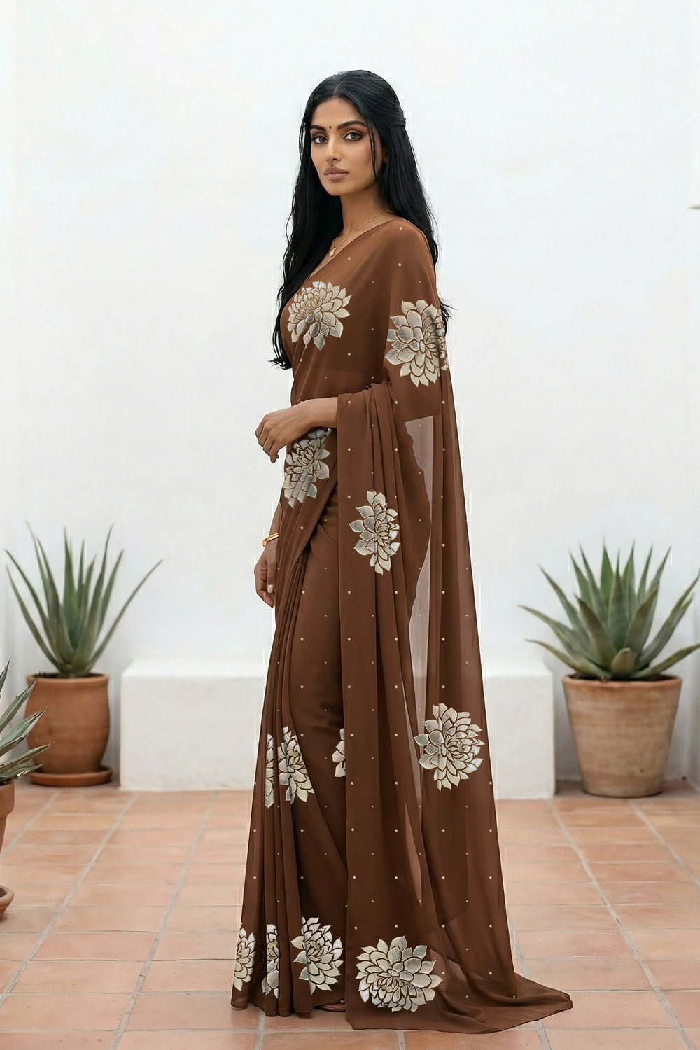 An Indian woman standing in a minimal white courtyard wearing a chocolate brown chiffon saree embellished with golden and sliver sequin of Dahlia flower motifs by Umaid Couture.  
