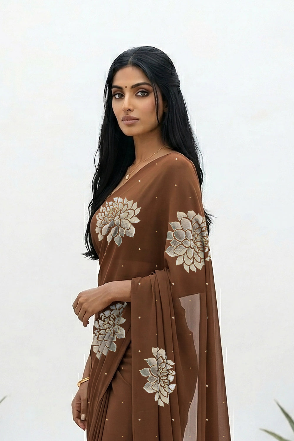 An Indian woman standing in a minimal white courtyard wearing a chocolate brown chiffon saree embellished with golden and sliver sequin of Dahlia flower motifs by Umaid Couture.  
