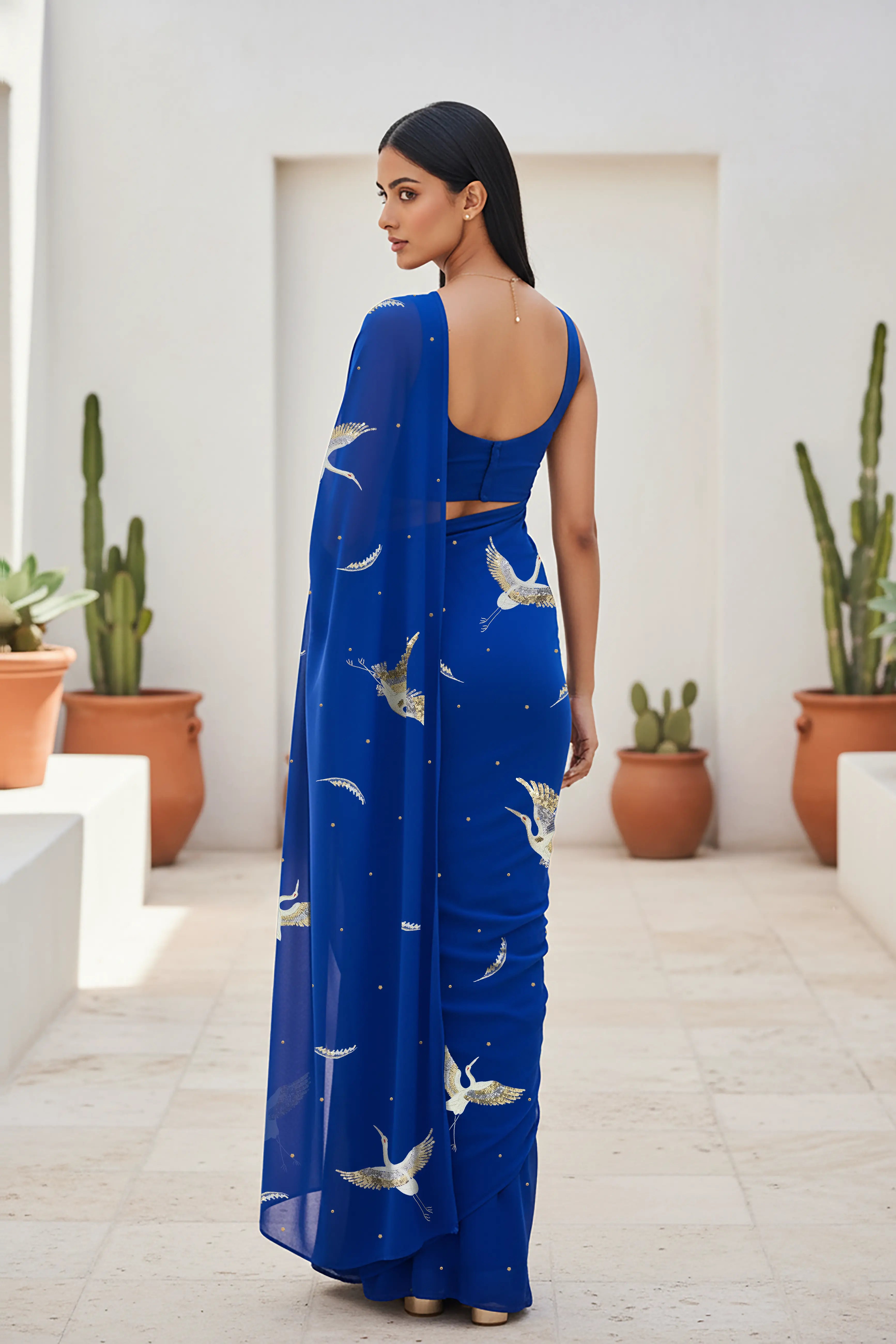 An Indian woman standing in a minimal Mexican courtyard wearing a revolutionary blue chiffon saree embellished with kurta bird motifs made with  shimmering Zari, Resham, Sequins, and Aari-Zardosi craft by Umaid Couture.  
