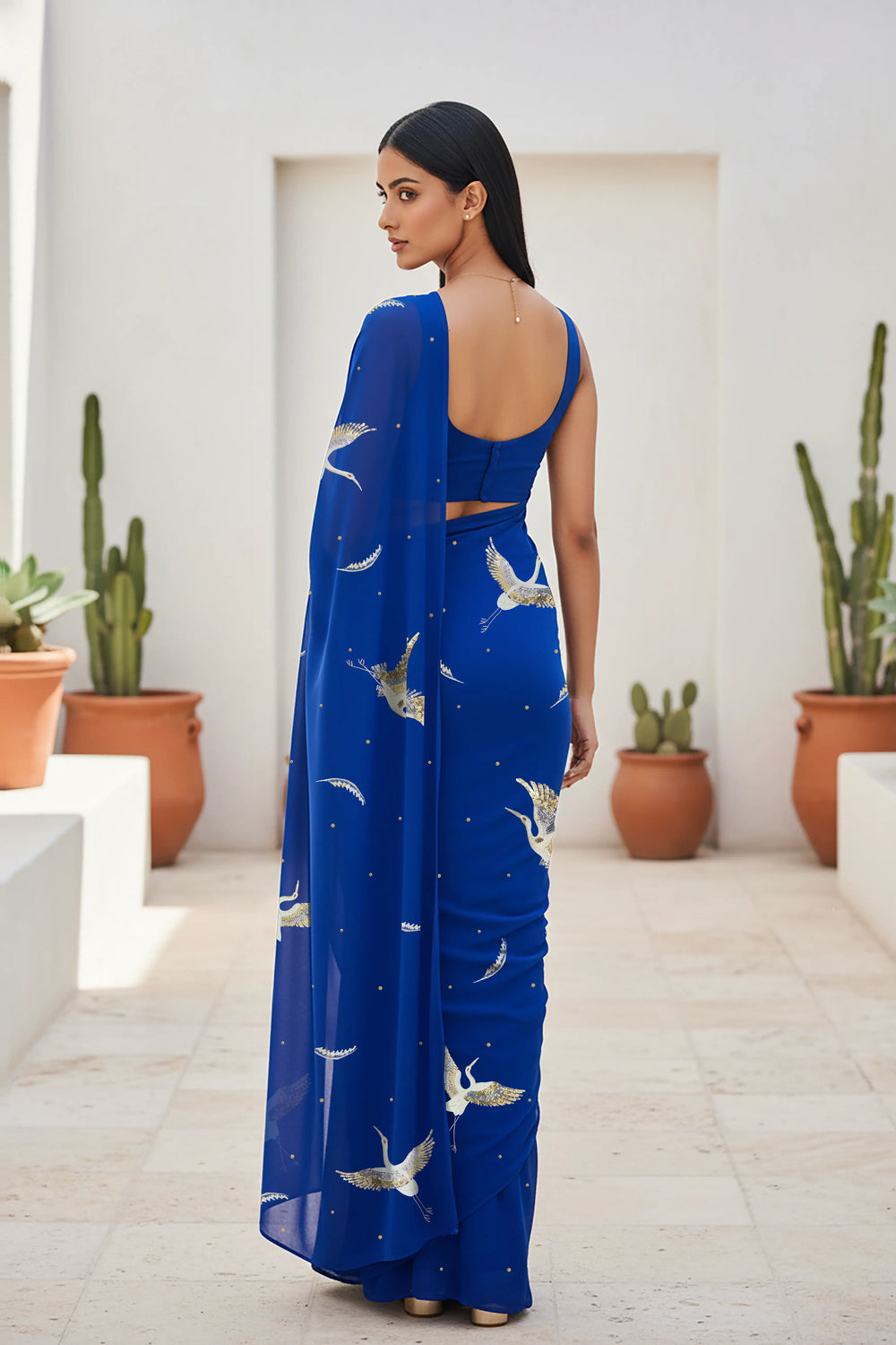 An Indian woman standing in a minimal Mexican courtyard wearing a revolutionary blue chiffon saree embellished with kurta bird motifs made with  shimmering Zari, Resham, Sequins, and Aari-Zardosi craft by Umaid Couture.  
