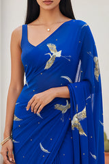 An Indian woman standing in a minimal Mexican courtyard wearing a revolutionary blue chiffon saree embellished with kurta bird motifs made with  shimmering Zari, Resham, Sequins, and Aari-Zardosi craft by Umaid Couture.  
