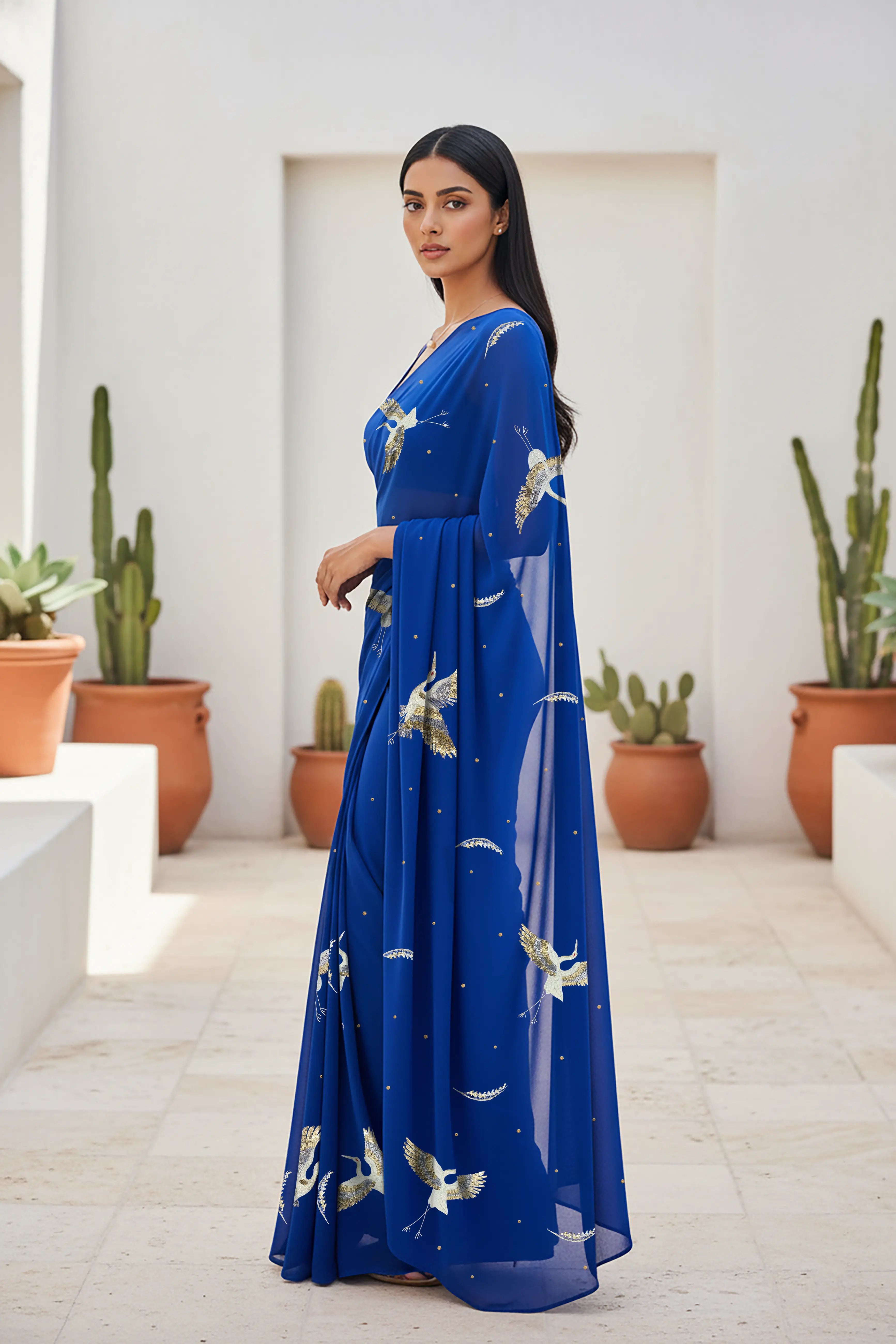 An Indian woman standing in a minimal Mexican courtyard wearing a revolutionary blue chiffon saree embellished with kurta bird motifs made with  shimmering Zari, Resham, Sequins, and Aari-Zardosi craft by Umaid Couture.  
