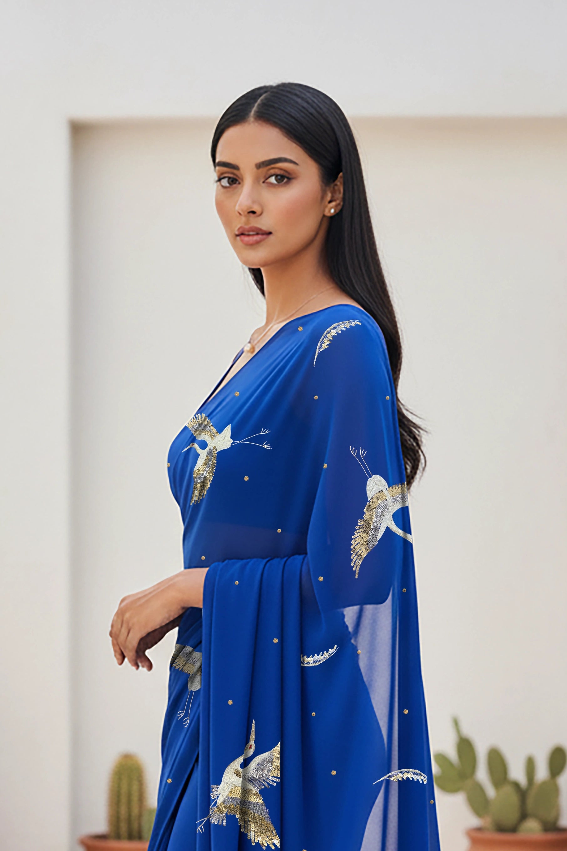 An Indian woman standing in a minimal Mexican courtyard wearing a revolutionary blue chiffon saree embellished with kurta bird motifs made with  shimmering Zari, Resham, Sequins, and Aari-Zardosi craft by Umaid Couture.  
