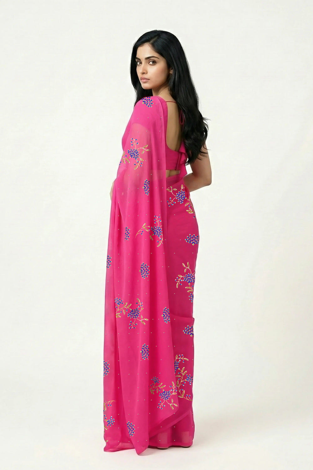 An Indian woman standing in a garden courtyard wearing a fuchsia pink chiffon saree embellished with blue and sliver sequin of lavender flower motifs by Umaid Couture.  
