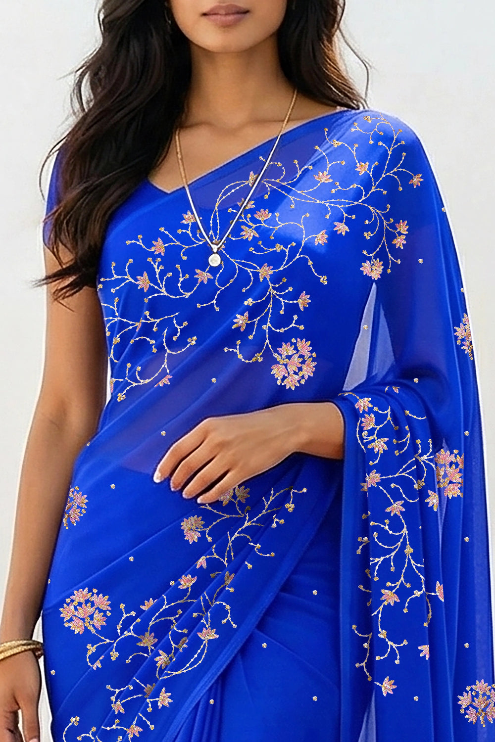 An Indian woman standing in a minimal white courtyard wearing a royal blue chiffon saree embellished with vgolden sliver and pink sequin vines of Stylized Jasmine by Umaid Couture.  
