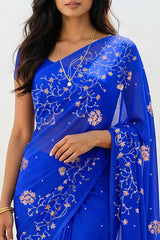 An Indian woman standing in a minimal white courtyard wearing a royal blue chiffon saree embellished with vgolden sliver and pink sequin vines of Stylized Jasmine by Umaid Couture.  
